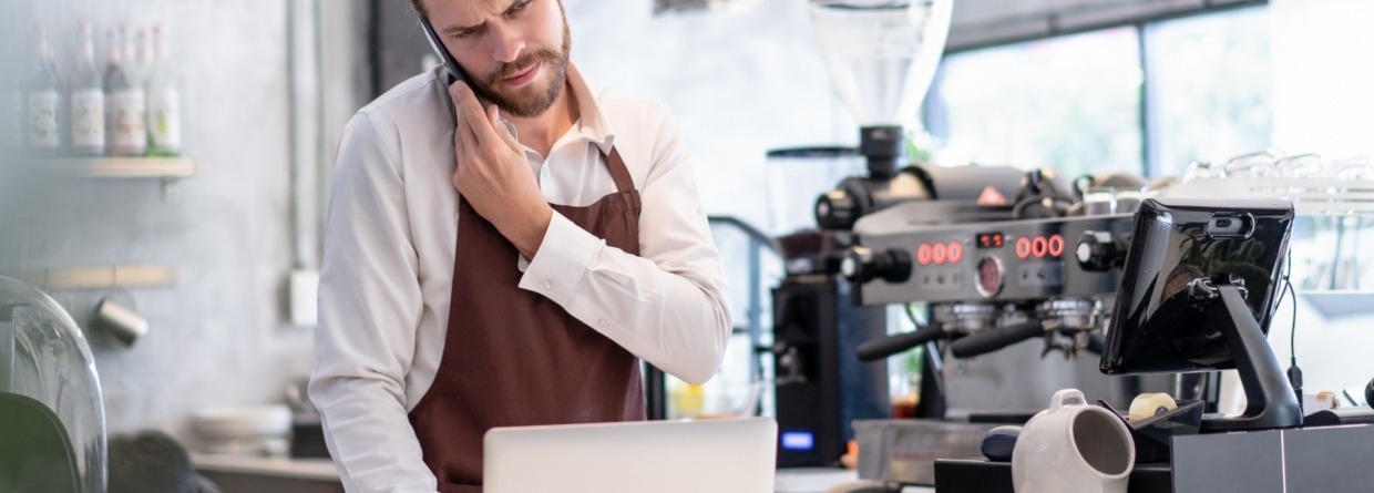 Serieuze bezorgde man wat een telefoongesprek voert en tussendoor naar informatie op zijn laptop aan het kijken is in een koffiebar